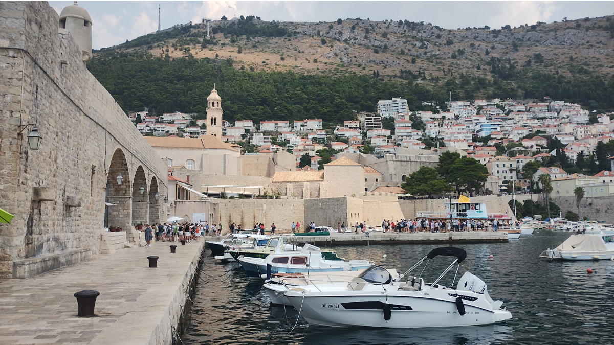 A scenic view of Dubrovnik, a historic coastal town with stone walls and arches along a waterfront promenade, small boats docked in the calm harbor, and hillside covered in white buildings with red roofs under a partly cloudy sky.