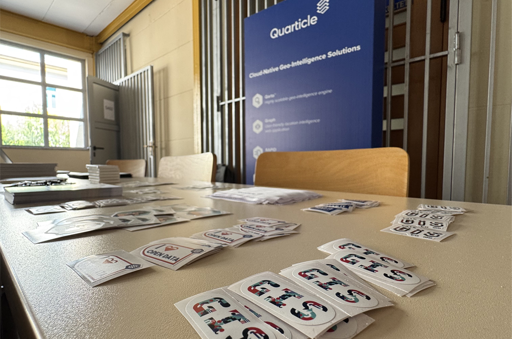 A table inside a room covered with various stacks of stickers and pamphlets. In the background, a blue vertical banner displays the Quarticle logo and the text 